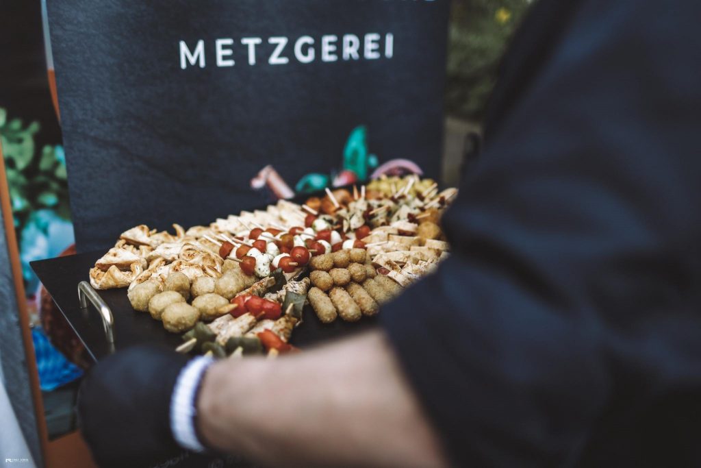 Eine Person traegt eine dekorative Wurstplatte vor einer Metzgerei.